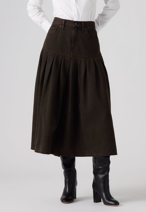 Femme portant une jupe midi plissée marron foncé taille haute, une chemise blanche et des bottes en cuir noires à talons montant jusqu'au genou.
