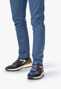 Sneaker con una combinazione di suede blu e pelle marrone, dotati di un logo bianco e suola nera testurizzata, abbinati a pantaloni blu aderenti.