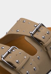 Μπεζ λουράκια σανδαλιών με ασημί τρουκς και ασημένιες μεταλλικές αγκράφες σε καφέ πάτο, κοντινή λήψη.