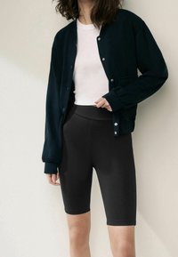 Giacca morbida blu navy con bottoni a pressione, camicia bianca a collo rotondo e pantaloni corti da biker neri a vita alta, creando un outfit monocromatico.