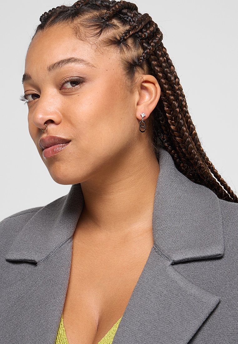 Cheveux tressés, manteau gris avec un col, portant des boucles d'oreilles pendantes avec une pierre claire et un détail en métal noir, présentant un design contemporain.