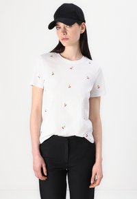 Witte t-shirt met korte mouwen en kleine geborduurde kersenpatronen, gecombineerd met zwarte broek en een zwarte baseballpet.