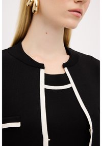 Svart stickad cardigan med vit kantning, med rund halsringning och dekorativa knappar, parat med en svart topp som har ett vitt rand.