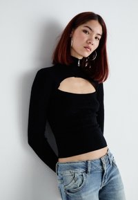 Black knit top with a high collar and cut-out under the neckline, featuring textured cable patterns. Paired with light blue denim jeans.