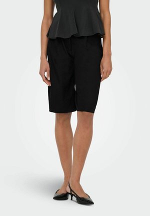 Kobieta ubrana w czarne, sięgające do kolan, dopasowane szorty, czarną bluzkę z baskinką oraz czarne sandały na niskim obcasie, stojąca na jednolitym tle.