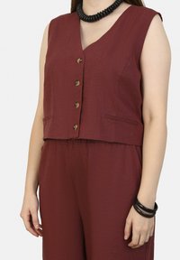 Bordeaux mouwloze vest met V-hals, voorzien van vijf knopen en twee zijzakken. Gemaakt boven een bijpassende hoge taillebroek.