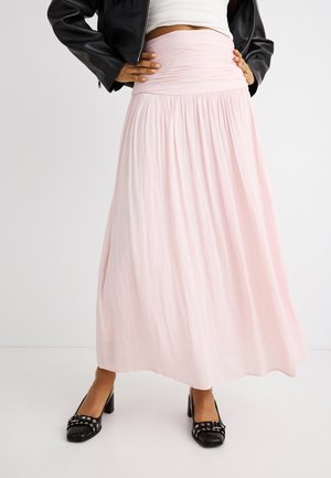 Kvinne iført en lys rosa plissert maxiskjørt, svarte blokkhælssko med nagler, hvit kort topp og svart skinnjakke, poserer med hendene på hoftene.