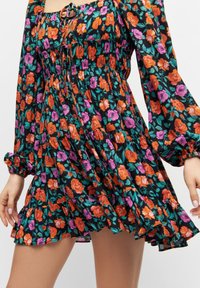 Robe fleurie avec un fond noir orné de roses orange et violettes. Manches longues, ourlet volanté et taille froncée avec un détail œillet.