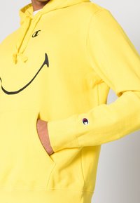 Sudadera amarilla de algodón, con un gran estampado de una cara sonriente negra, bolsillo tipo canguro y pequeño logo bordado en el puño.