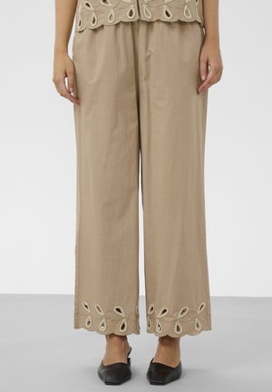 Pantalon beige à jambes larges avec ourlet festonné brodé, porté avec des chaussures plates noires à bout pointu.