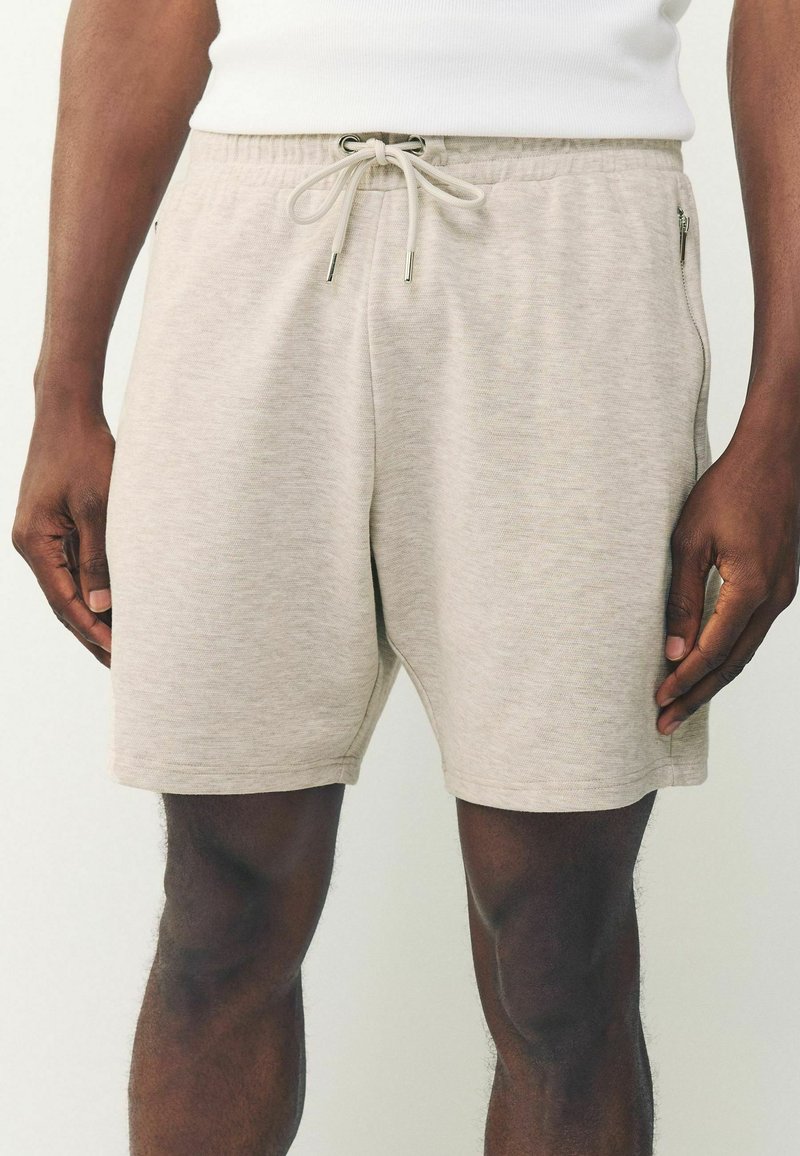 Hombre vistiendo pantalones cortos beis claros con cordón y bolsillo lateral con cremallera, acompañado de una camiseta sin mangas blanca, contra un fondo liso.