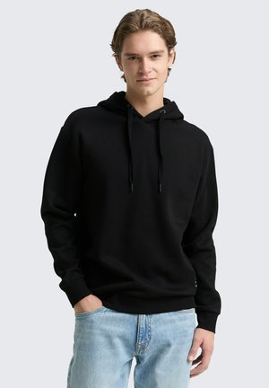 Junger Mann trägt einen schwarzen Kapuzenpullover mit Kordeln und hellblauen Jeans, steht mit einer Hand in der Tasche.