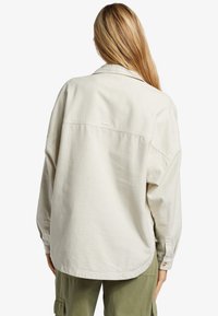 Cream-colored oversized shirt with a soft texture, pointed collar, and buttoned cuffs, featuring a rounded hem and seam detailing at the back.
