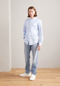 Camicia a maniche lunghe azzurra con un sottile motivo a foglie bianche, design con bottoni, abbinata a jeans azzurri e sneakers bianche.