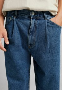 Denim jeans with a dark blue wash, featuring a high waist, pleats at the front, and visible stitching. Worn with a light-colored top.