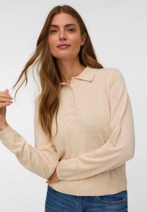 Femme avec de longs cheveux châtain clair portant un polo beige à manches longues et un jean bleu, tenant délicatement une mèche de ses cheveux.