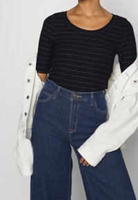 Černý pruhovaný top s krátkými rukávy, spárovaný s vysokými modrými džíny. Bílá denimová bunda leží přes ruku.