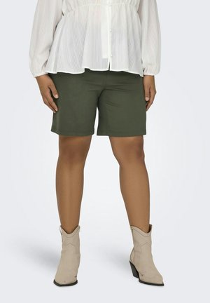 Nedre del av en person som har på seg olivengrønne knelange shorts, hvit bluse med lange ermer, og beige ankelstøvletter med hæl på hvit bakgrunn.