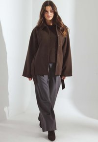 Hnedá oversized košeľa s gombíkmi, pod ňou textúrovaný pletený sveter, skombinované s tmavosivými širokými nohavicami a hnedými členkovými čižmami, neutrálne pozadie.