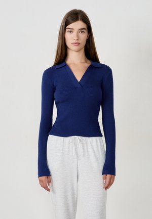 Junge Frau mit langen braunen Haaren, die einen enganliegenden blauen Rippen-V-Ausschnitt-Pullover und hellgraue Kordelzughosen trägt, vor einem einfarbigen Hintergrund.