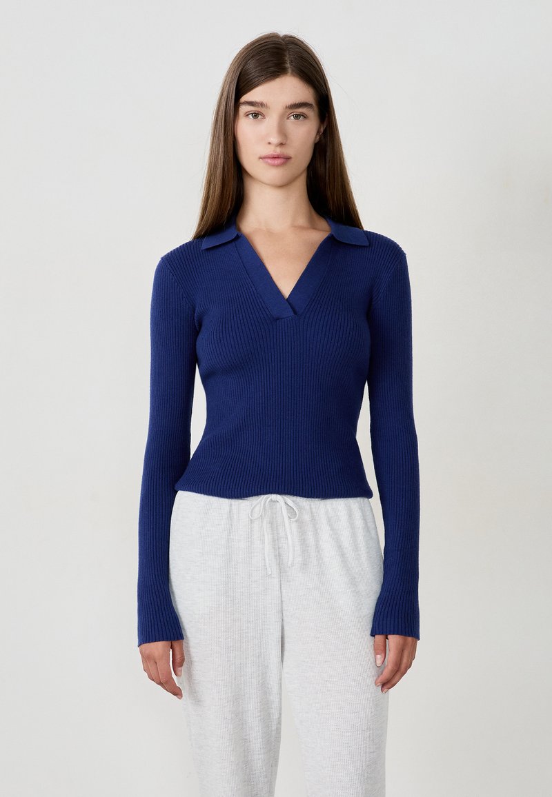 Młoda kobieta z długimi brązowymi włosami, ubrana w dopasowany niebieski sweter w prążki z dekoltem w serek i jasnoszare spodnie z troczkami na tle jednolitego tła.