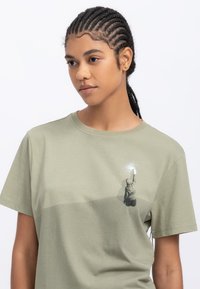 T-shirt in cotone verde oliva con maniche corte, caratterizzato da una stampa grafica di una figura che si allunga verso l'alto sul lato sinistro. Texture liscia.
