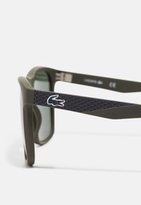 Óculos de sol de plástico verde com lentes escuras. Apresenta uma haste preta texturizada com um padrão hexagonal e um detalhe do logótipo.