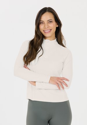 Top de manga larga color beige claro con cuello de media cremallera y diseño ajustado. Tejido suave y elástico con un ajuste cómodo. Combinado con leggings verde oscuro.