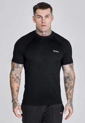 Mężczyzna z tatuażami ubrany w dopasowaną czarną koszulkę Sik Silk i czarne spodnie, stojący przodem na neutralnym tle.