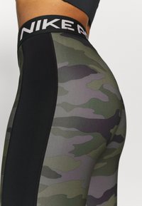 Nike leggings hebben een zwarte elastische tailleband met het logo. Het lichaam heeft een camouflagepatroon in tinten groen en grijs.