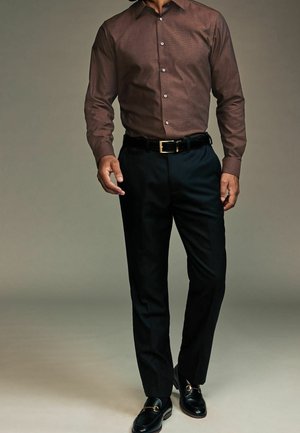 Homme portant une chemise marron foncé boutonnée, une ceinture noire, un pantalon de costume noir et des mocassins noirs avec un détail de boucle dorée.