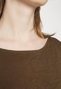 Ruskea kulho-kauluksinen paita, jossa on teksturoitu, kevyt kangas. Kaula-aukko istuu lähellä solisluita ja siinä on hieman ribattu reunus.