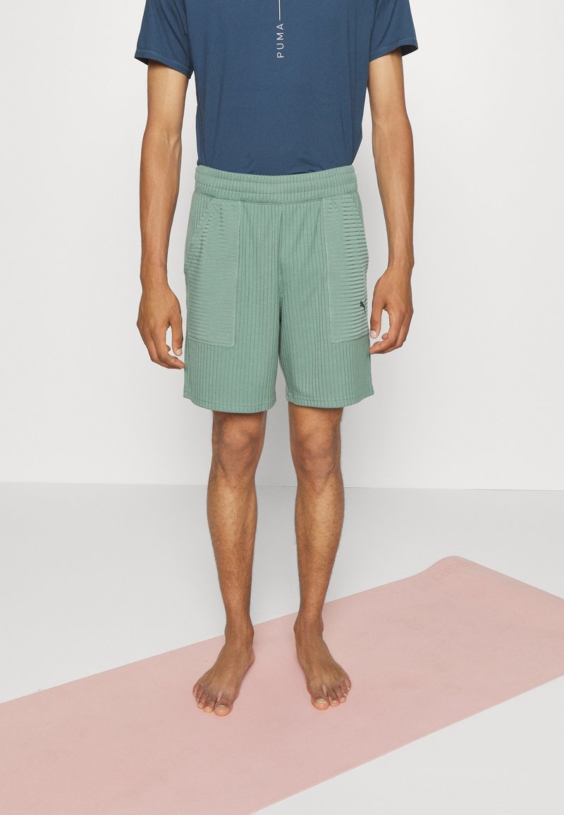 Zelené pruhované šortky s dvěma předními kapsami, elastickým pasem a malým logem. Nosit s námořnickomodrým tričkem. Naboso na růžové podložce.