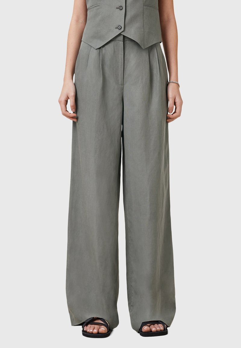 Femme portant un pantalon large gris ample à taille haute avec un gilet assorti et des sandales noires ouvertes aux orteils, sur un fond uni.