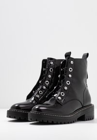 Černé lakované kozačky kotníkové s robustní podrážkou, obsahující velké stříbrné oči, šňůrky a poutko na zadní straně.