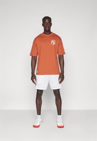 Rood-oranje t-shirt met een wit NY-logo, gecombineerd met witte sportshorts en grijze sneakers met rode accenten. Eenvoudig, casual ontwerp.