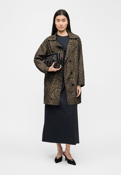 Γυναίκα που φοράει παλτό με λεοπάρ σχέδιο πάνω από μαύρο φόρεμα, κρατώντας μαύρο τσαντάκι με υφή, και φοράει μαύρες μυτερές γόβες, στέκεται μπροστά σε λευκό φόντο.