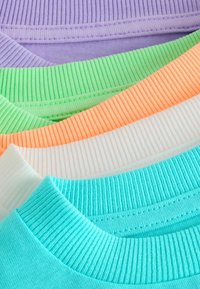 Pile de pulls en tricot côtelé pliés dans des tons pastel violet, vert, orange, blanc et bleu, montrant les détails du col et de la texture.