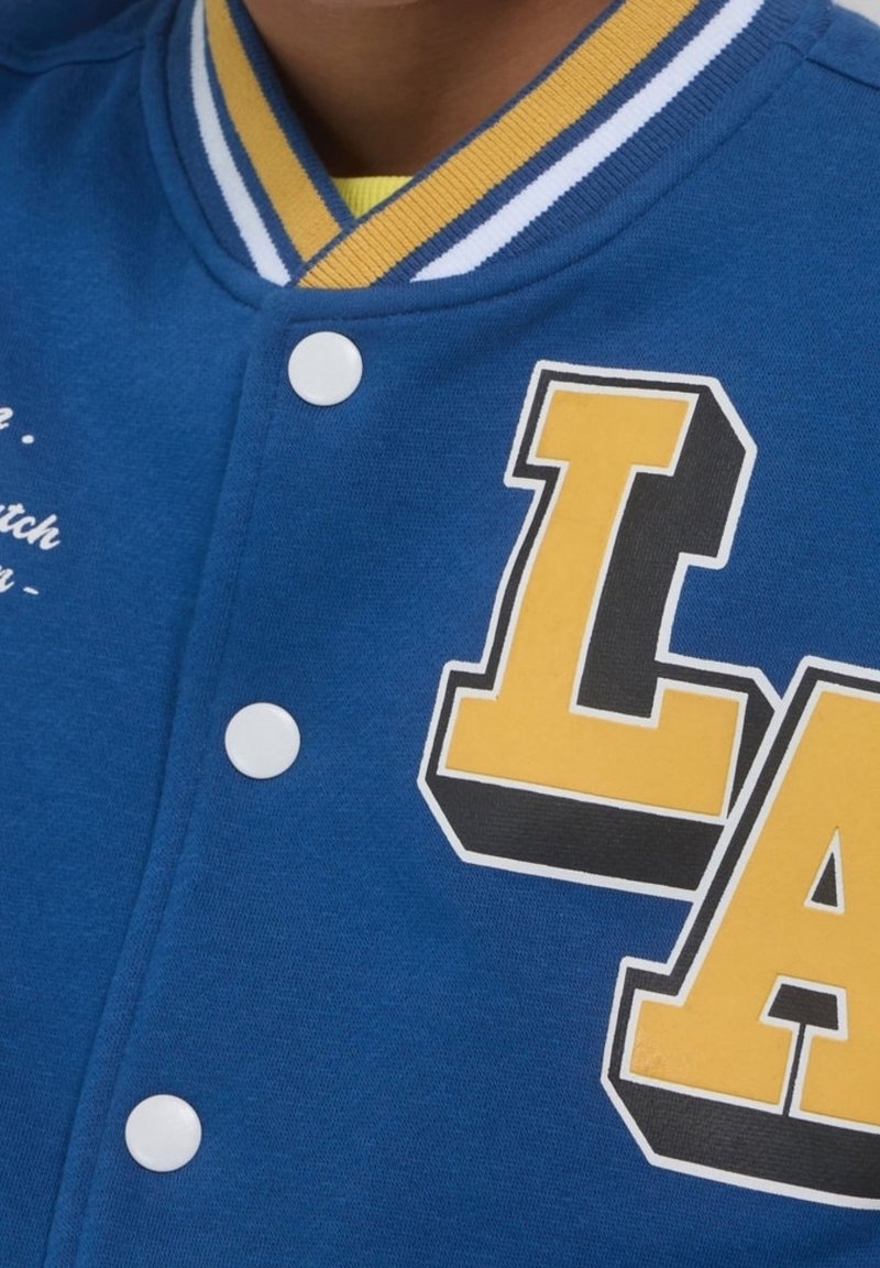 Niebieska kurtka varsity z żółtym logo "LA" i białymi guzikami. Posiada kołnierz w paski w kolorach niebieskim, żółtym i białym. Na tkaninie znajduje się tekst.