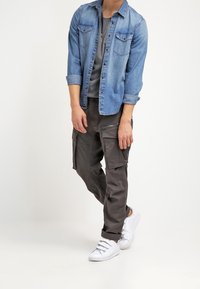 Chemise en denim bleu clair avec deux poches poitrine, t-shirt gris en dessous, pantalon cargo gris foncé avec poches latérales, et baskets blanches.