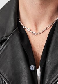 Primo piano di una persona che indossa una collana di catena d'argento con maglie irregolari, una camicia nera abbottonata e una giacca di pelle nera.