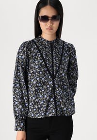 Floral-patterned blouse with black background, light blue and green flowers, high neckline, puffed sleeves, and contrast trim detail.