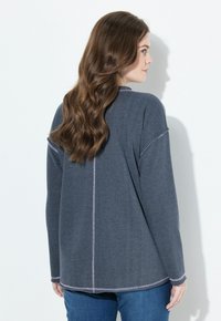 Donna con lunghi capelli castani, che indossa un maglione grigio scuro largo con cuciture visibili e jeans blu, voltata di lato contro un muro bianco.