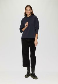 Donkergrijze hoodie met lange mouwen, gecombineerd met zwarte wijde broek en zwarte enkellaarzen met groene zolen. Hete stof.