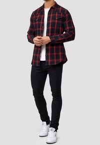 Mies, joka on pukeutunut punamustaraidalliseen paitaan valkoisen t-paidan päälle, mustiin skinny-farkkuihin ja valkoisiin Reebok-tennarisiin, seisoo yksinkertaista taustaa vasten.