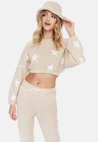 Béžová cropped mikina s bielymi hviezdami, skombinovaná s rovnakou teplákovou súpravou. Model nosí huňatý béžový klobúk s širokou krempou a usmieva sa.