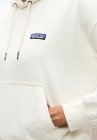 Άτομο που φοράει μια εξωλεκάνη Patagonia σε εκρού χρώμα, με το χέρι στην μπροστινή τσέπη και εμφανείς κορδόνια να κρέμονται από τη κουκούλα.