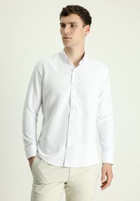 Camicia bianca a maniche lunghe con bottone, dal tessuto morbido e colletto piccolo. Presenta bottoni bianchi e orlo leggermente curvo.