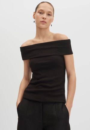 Donna che indossa un top nero scollato sulle spalle e pantaloni neri, in piedi con le mani nelle tasche davanti a uno sfondo chiaro e uniforme.