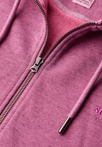 Hoodie zippé en tissu rose clair, avec une fermeture éclair en métal, des cordons de serrage et des détails brodés. Texture douce avec une finition lisse.